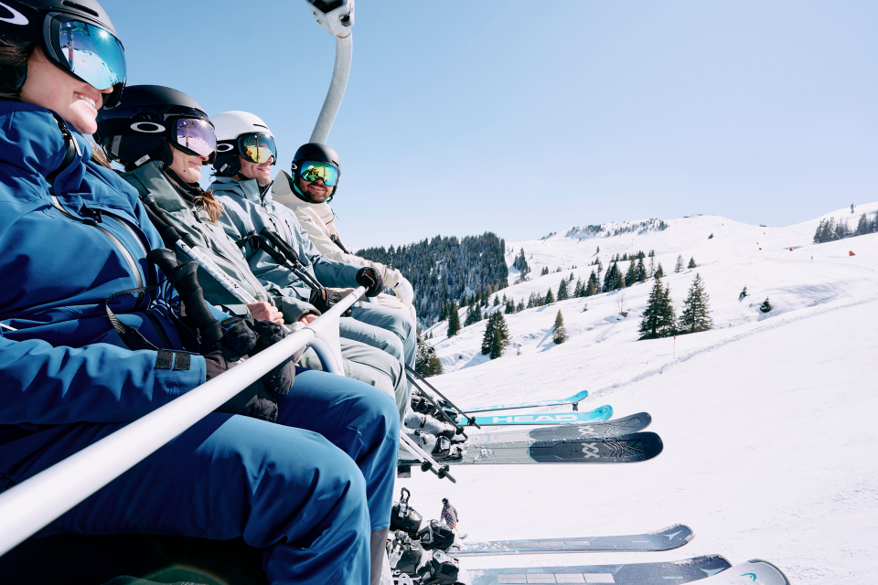 Four skiers are sitting in a chairlift, with a white ski slope visible in the background.
