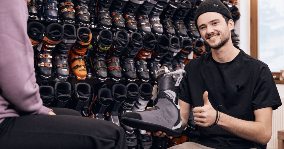 Mitarbeiter hält den Innenschuh eines Skischuhs in der Hand, lächelt und zeigt mit dem Daumen nach oben. Im Hintergrund sieht man eine Regal voller Skischuhe.