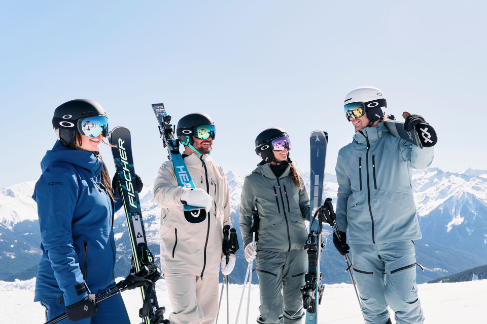 Four skiers are standing in the snow, smiling and looking at each other.